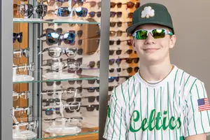 A young man wearing sunglasses and a Boston Celtics baseball cap stands in front of a glass cabinet filled with sunglasses.