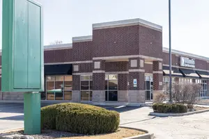 An exterior view of a storefront featuring a brick facade, large glass windows, and a green sign on the left side.