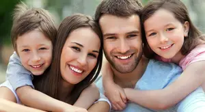 A family of four, including a man, a woman, and two children, are smiling and posing for a photograph in a park, with a blurry background of trees and plants