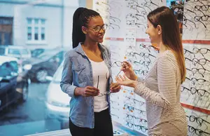 Two women discussing glasses in an eyewear store, with a wall display of various frames.