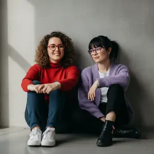 Two women sitting on the floor in front of a wall with a shadow cast on it, wearing glasses and smiling at the camera.