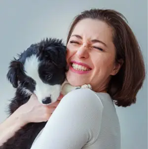 A woman with brown hair is holding a black and white dog and smiling with closed eyes