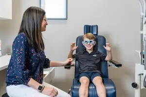 A young boy wearing goggles is sitting in a chair while a woman with glasses holds his hands.