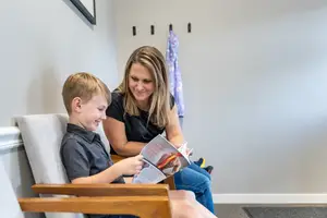 A woman is sitting on a couch with a boy and they are reading a book together