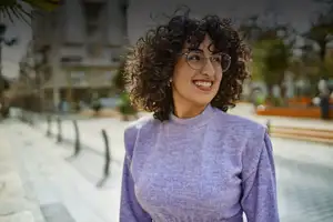Smiling person with curly hair and glasses, wearing a purple sweater, standing outdoors in a city setting.