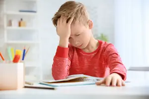 A young boy sitting at a desk with his hand on his forehead, looking frustrated while studying.