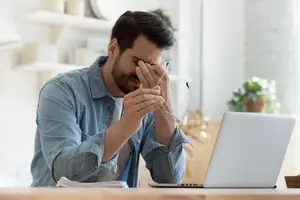 A man covering his eyes with his hand while sitting at a desk with a laptop in front of him.