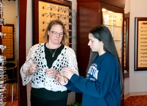 An adult woman and a young girl are standing in front of a display case of eyeglasses in a store.