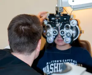 A man adjusts a woman's eyeglass while she looks through an eye chart in an exam room