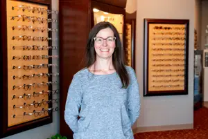 A woman is standing in front of a wall with framed glasses on display, smiling and wearing glasses.