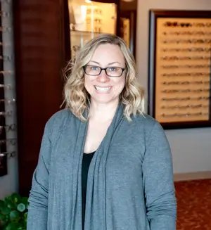 A woman with blonde hair wearing glasses and a gray cardigan stands in a room with a plant and a wall with a display case behind her.