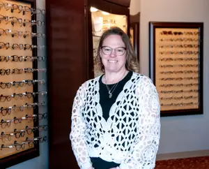 A woman wearing glasses and a necklace stands in front of a wall with a display of eyeglasses and framed artwork.