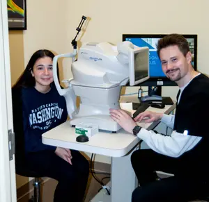 A smiling girl and a man are sitting next to each other, the man is using an eye exam machine while the girl is sitting in front of a monitor with a Dell logo