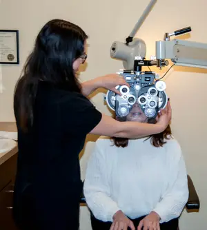 A woman is adjusting an eye examination machine on a seated patient in an office setting.