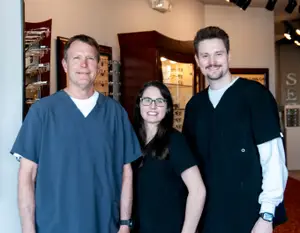 Three people are standing in a room and smiling at the camera. The man on the left is wearing a blue shirt, the woman in the middle is wearing a black shirt and glasses, and the man on the right is wearing a black shirt and a watch. Behind them is a wooden cabinet with a glass door, a shelf with a display of glasses, and a wall with a mirror and a lamp.