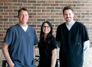 Three individuals, two men and one woman, are standing together in front of a brick wall, dressed in scrubs and smiling for the camera.