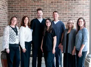Seven people standing in front of a brick wall wearing different colored shirts and pants, with two men wearing lab coats, and a woman in the center wearing a necklace