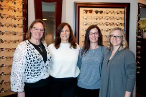 Four women wearing glasses are standing close to each other in front of a display of glasses in an optical shop.