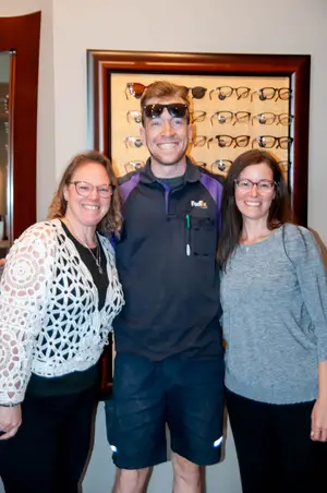 Three people stand next to each other in front of a wall with a picture frame of glasses.