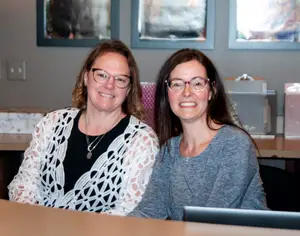 Two women wearing glasses and smiling are sitting on chairs in front of a desk. The woman on the left is wearing a necklace and a bracelet. The woman on the right is wearing a necklace and a ring. Behind them are some frames on the wall and some boxes on the desk.