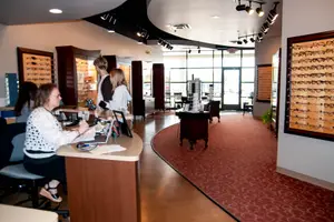 Inside a store with people standing and sitting near a desk, a woman sitting at a desk, a cabinet, and glass display cases with many glasses on the wall