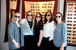 Five women wearing sunglasses are standing in front of a wall with frames displayed on it.