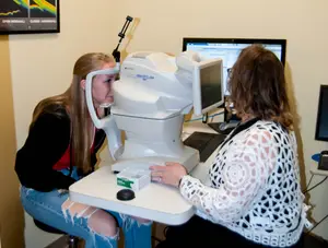 A young woman has her eyes examined by an older woman using a medical eye scanner in a clinical setting.