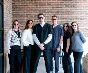 Six people wearing glasses stand in front of a brick wall, with a man in a black shirt in the middle, and a man on the right wearing a blue shirt and sunglasses.
