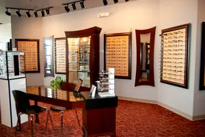 An optical shop with a desk, chairs, and glass cabinets displaying eyeglasses, with a mirror and lights on the wall, and a potted plant in the corner.