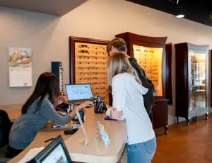 Three people in an optometrist's office, one of them working at a desk with a computer and glasses on display.