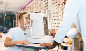 A young boy is having his eyes examined by an optician in a white coat in a well-lit store with shelves of glasses and equipment.