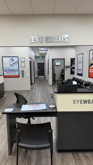 An eye exam room with a desk, chairs, and a sign saying Eye Exams. Behind it is a hallway with a door and a sign. On the right side is a desk with eyeglasses and a basket. On the left is a wall with a poster and a sign. On the ceiling are lights. In the distance is another room with a desk and a chair.
