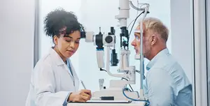 A man and a woman in a lab, with the woman looking through an eye exam machine and holding a pen and paper while the man is looking at her and holding a headband
