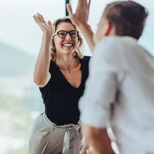A woman wearing glasses and a necklace is smiling while raising her hands in front of a man in a white shirt who is raising his hands as well in an indoor area