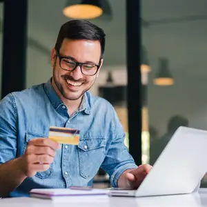 A man is smiling and holding a credit card while looking at a laptop