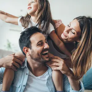 A family of three, with a man, woman, and young girl, is sitting on a couch, smiling and laughing together, with the man holding the girl on his shoulders.