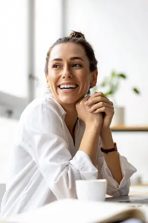A woman sitting at a desk and smiling with a cup on the table in front of her.