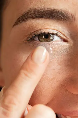 A close-up of a woman's face, she is wearing contact lenses and is about to put another one on her eye.