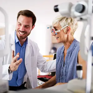 An older woman wearing glasses is smiling while talking to a man in a white lab coat.