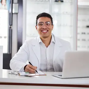 Man in a lab coat sitting at a desk with a laptop and a pen, smiling and looking at something