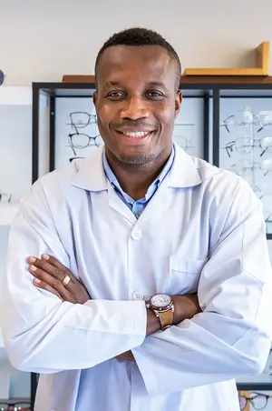 A smiling man in a white lab coat and watch stands with arms crossed in front of a shelf of glasses