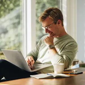 A man is sitting on a chair in front of a desk with a laptop, a cup, a book, a pen, and a cell phone. He is wearing eyeglasses, a watch, and a green sweater. He is smiling and seems to be posing for a photo. Behind him is a glass window with a view of trees and plants.