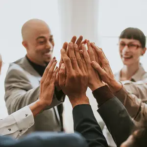A group of people are standing in a room, and they are holding their hands together and smiling, probably in a meeting room.
