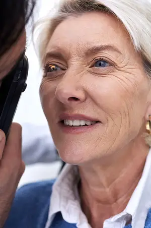 An elderly woman with white hair is having her eye examined by a doctor wearing a white coat and holding a black cell phone.