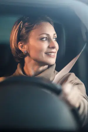 A smiling woman wearing a brown coat and earrings drives a car while holding the steering wheel with her left hand and her seatbelt fastened