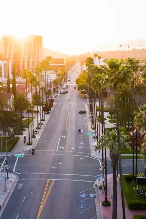 A road with tall palm trees and green street lights on either side. There are also buildings and a mountain in the distance. The sun is setting behind the mountain, casting a warm glow on the road and the surroundings. A car is driving on the road, and there are also some people walking on the sidewalk.