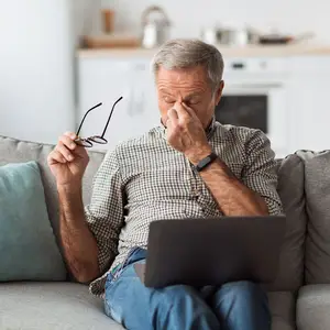 An older man sitting on a couch, holding a pair of glasses, and covering his nose with his hand. He seems to be feeling sick.