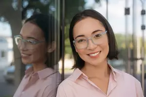 A woman in glasses is smiling and looking at her reflection in a glass wall.