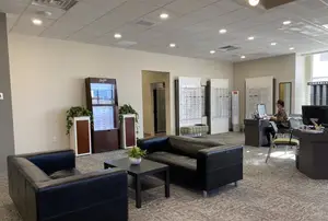 Interior of a business office with seating and a desk, a woman sitting at the desk, and a monitor, a potted plant, and an eyewear display case in the background.