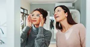 Two women standing in front of a mirror at an eyeglass store, one woman is trying on glasses and the other is laughing